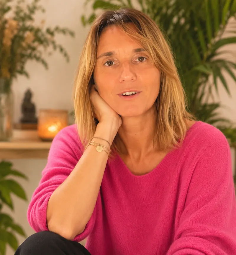 Caroline Senges, souriante, en pull rose, la main contre la joue, devant des plantes et une bougie allumée.