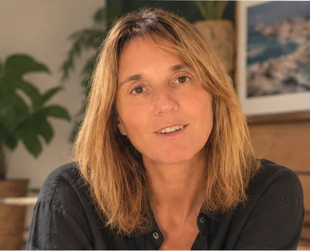 Caroline Senges en chemise noire, en train de parler, devant des plantes et une photo de bord de mer.